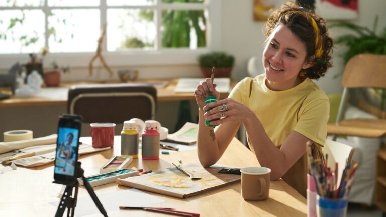 A young woman engaged in painting while being recorded on a mobile phone placed on a tripod